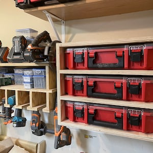 May include: A wall-mounted storage system for tools and small parts. The system features a wooden shelf with multiple compartments for storing small items, and a separate section with hooks for hanging tools. The system also includes three red plastic storage boxes with clear lids.