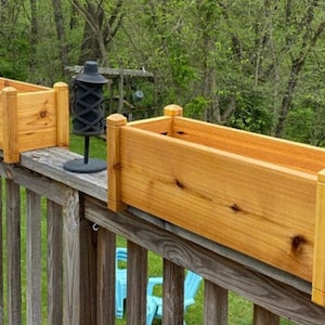 May include: Two wooden planter boxes are mounted on a wooden railing. The boxes are made of natural cedar wood and have a light brown color. The boxes are rectangular in shape and have a slightly rounded edge. The boxes are empty and ready to be planted with flowers or herbs.