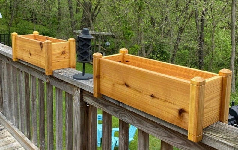 May include: Two wooden planter boxes are mounted on a wooden railing. The boxes are made of natural cedar wood and have a light brown color. The boxes are rectangular in shape and have a slightly rounded edge. The boxes are empty and ready to be planted with flowers or herbs.