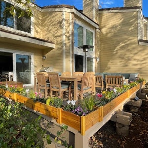 May include: A wooden patio with a dining table and chairs, surrounded by raised flower beds filled with blooming flowers. The patio is attached to a yellow house with large windows.
