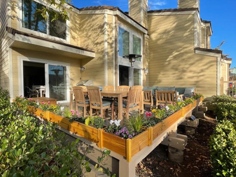 May include: A wooden patio with a dining table and chairs, surrounded by raised flower beds filled with blooming flowers. The patio is attached to a yellow house with large windows.