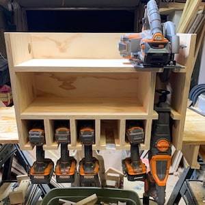 May include: A wooden wall-mounted storage shelf with six compartments for tools. The shelf is holding five orange and black cordless drills and a black and orange reciprocating saw. The shelf is also holding a black and orange circular saw on the top shelf.