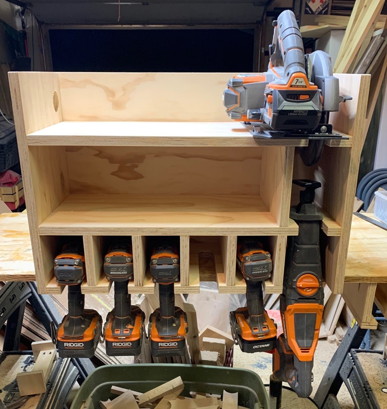 May include: A wooden wall-mounted storage shelf with six compartments for tools. The shelf is holding five orange and black cordless drills and a black and orange reciprocating saw. The shelf is also holding a black and orange circular saw on the top shelf.