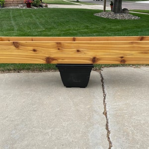 May include: A long, rectangular wooden planter box made of light brown wood, supported by a black plastic pot. The planter box is empty and sits on a concrete surface, with green grass and a house in the background.
