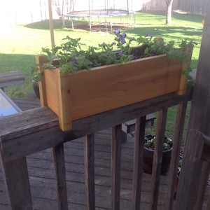 May include: A wooden planter box with purple flowers growing in it. The planter is mounted on a wooden railing on a deck.