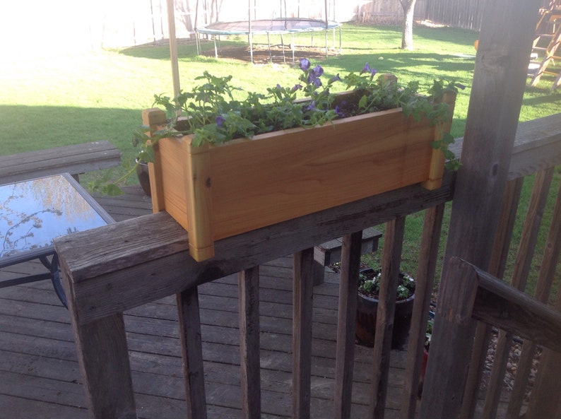 May include: A wooden planter box with purple flowers growing in it. The planter is mounted on a wooden railing on a deck.
