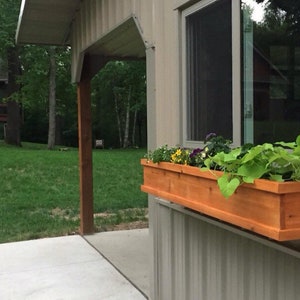 May include: A wooden window box planter with a variety of green plants and flowers. The planter is mounted on the exterior wall of a building.