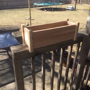 May include: A wooden planter box, made of natural cedar wood, is mounted on a wooden railing. The planter box is empty and ready for planting.