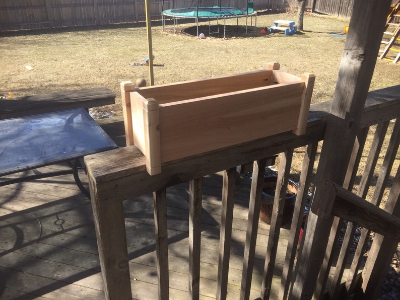 May include: A wooden planter box, made of natural cedar wood, is mounted on a wooden railing. The planter box is empty and ready for planting.