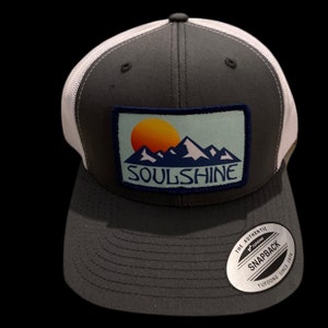 May include: A gray and white trucker hat with a patch that says "SOULSHINE" and features a mountain range and a setting sun. The hat has a black mesh back and a white snap closure. The hat also has a white sticker on the back that says "THE AUTHENTIC YUPOONG SNAPBACK SINCE 1974."