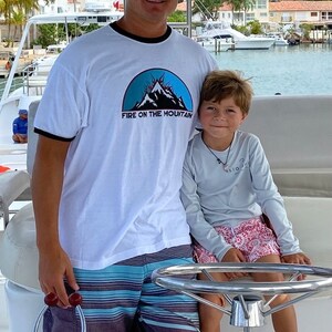 Puede incluir: Un hombre y un ni&ntilde;o est&aacute;n en un barco. El hombre lleva una camiseta blanca con un gr&aacute;fico de monta&ntilde;a azul y el texto "FIRE ON THE MOUNTAIN". Tambi&eacute;n lleva una gorra de b&eacute;isbol gris con la palabra "PAPPY" en ella. El ni&ntilde;o lleva una camisa de manga larga azul claro con un dise&ntilde;o rojo y blanco. El hombre est&aacute; sujetando el volante del barco.