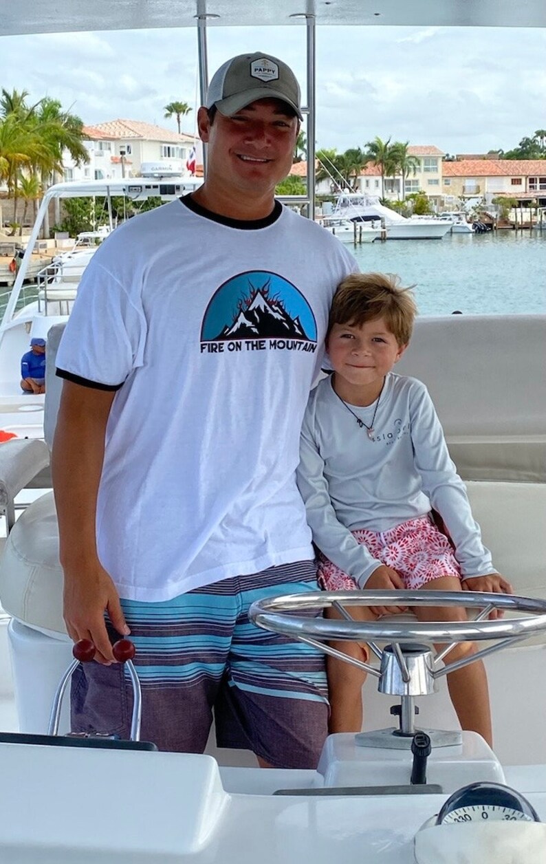 Puede incluir: Un hombre y un ni&ntilde;o est&aacute;n en un barco. El hombre lleva una camiseta blanca con un gr&aacute;fico de monta&ntilde;a azul y el texto "FIRE ON THE MOUNTAIN". Tambi&eacute;n lleva una gorra de b&eacute;isbol gris con la palabra "PAPPY" en ella. El ni&ntilde;o lleva una camisa de manga larga azul claro con un dise&ntilde;o rojo y blanco. El hombre est&aacute; sujetando el volante del barco.