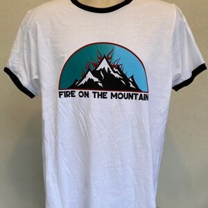 Puede incluir: Camiseta blanca con cuello de canal&eacute; negro. La camiseta presenta un gr&aacute;fico de monta&ntilde;a azul, azul verdoso y negro con llamas rojas. El texto "Fire on the Mountain" est&aacute; impreso debajo del gr&aacute;fico.