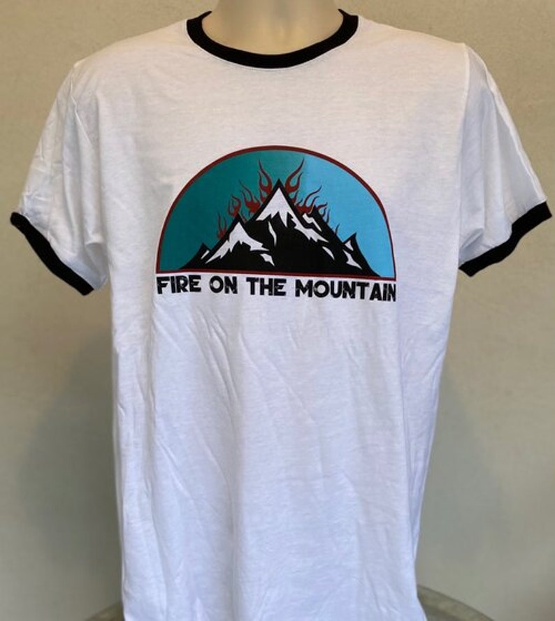 Puede incluir: Camiseta blanca con cuello de canal&eacute; negro. La camiseta presenta un gr&aacute;fico de monta&ntilde;a azul, azul verdoso y negro con llamas rojas. El texto "Fire on the Mountain" est&aacute; impreso debajo del gr&aacute;fico.