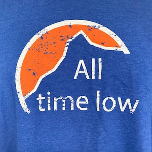 May include: A blue t-shirt with a distressed orange graphic of a mountain peak and the text "All time low" in white.