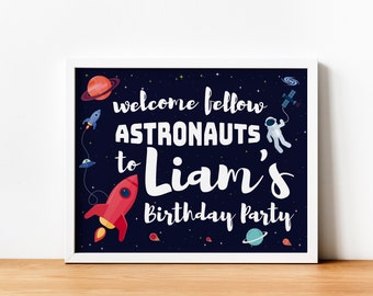 May include: A blue and black space-themed birthday party sign with white text that reads "Welcome fellow astronauts to Liam's Birthday Party". The sign features a rocket, a spaceship, an astronaut, and planets.
