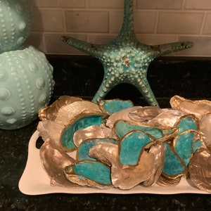 May include: A display of decorative oyster shells with gold edging and turquoise interiors, presented on a white tray. A turquoise starfish and two light blue spheres are in the background, evoking a coastal aesthetic.