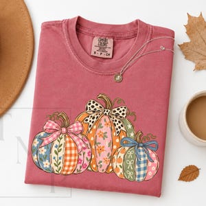 May include: A dusty rose short-sleeve t-shirt featuring a graphic of three pumpkins with various patterns including gingham, floral, and polka dots, each adorned with a bow. The shirt has a Comfort Colors label at the neck and a crew neckline.