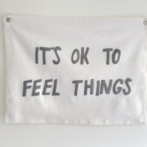 Può includere: Striscione in tela bianca con il testo "It's ok to feel things" stampato in nero.