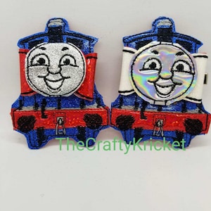 May include: Two blue and red train engine character hair clips with a white bow and a silver glitter face. The clip on the left has a silver glitter face and the clip on the right has a holographic face.
