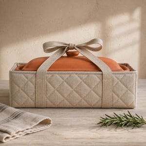 May include: A rectangular terracotta baking dish with a matching lid, encased in a beige quilted fabric carrier. The carrier features a diamond-stitched pattern and is secured at the top with a tied fabric bow made from the same beige material.