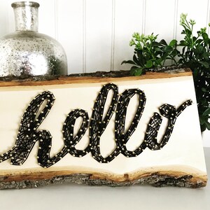 May include: A wooden sign with the word "hello" spelled out in black string wrapped around gold tacks. The sign is on a white surface with a silver vase and green plants in the background.
