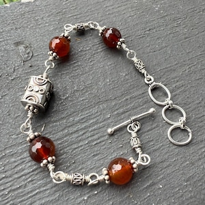 May include: Silver bracelet featuring faceted, reddish-brown beads and a decorative cylindrical charm. The bracelet has a toggle clasp and is composed of interconnected links. A piece of jewelry.
