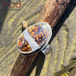 May include: A silver ring with an oval-shaped, multi-colored stone. The stone features orange, brown, and blue patterns. The ring band has a decorative design. The ring is displayed on a wooden stick.