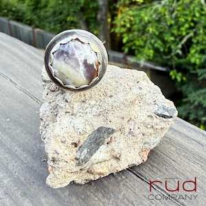 May include: A silver ring with a large, round, multi-colored stone. The stone has shades of white, brown, and purple. The ring is set on a piece of rough, textured concrete. The word "rud" and "COMPANY" are visible.