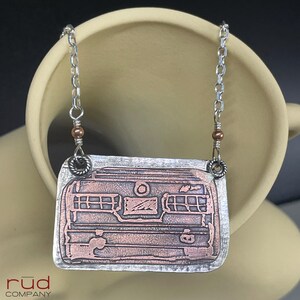 May include: A silver-toned necklace with a rectangular pendant featuring a copper-colored design of a vintage camper. The pendant has etched details of windows and other camper features. The necklace chain is silver with small copper-colored beads.