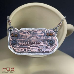 May include: A handcrafted silver necklace with a copper-toned pendant. The pendant features a detailed design of a vintage train, with amber and blue-green gemstone accents. The necklace is suspended from a delicate silver chain.