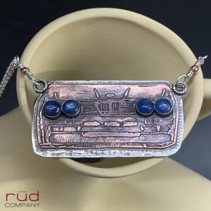 May include: A handcrafted necklace featuring a rectangular pendant with a copper and silver design. The pendant has three pairs of blue gemstone accents. The necklace is suspended from a silver chain.