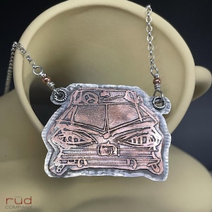 May include: A copper-colored pendant necklace featuring a detailed car design. The pendant is framed in silver and hangs from a delicate silver chain. The car design includes headlights, a steering wheel, and other intricate details.