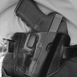 May include: A black leather holster with a black handgun inside. The holster is worn on a person's waist and is secured with a belt.