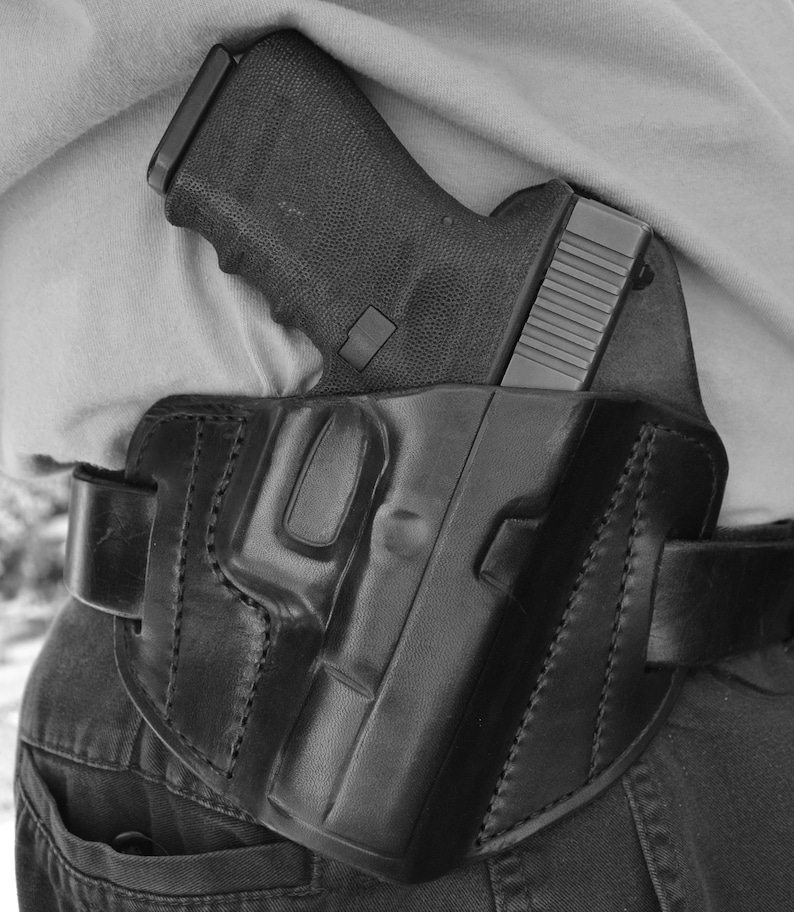 May include: A black leather holster with a black handgun inside. The holster is worn on a person's waist and is secured with a belt.