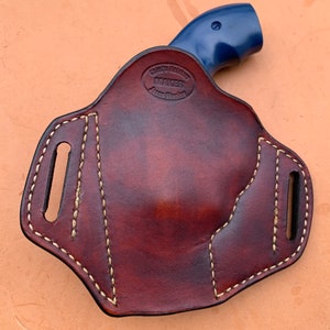 May include: A brown leather holster with a white stitch design and a blue gun inside. The holster has two belt loops and the text "Black's Holster Maker Jason Brown" is embossed on the back.