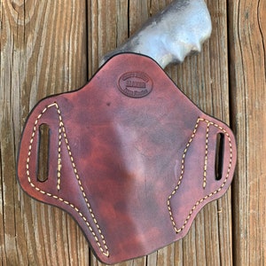 May include: A brown leather holster with a light brown stitching detail. The holster has a cut-out for a gun and a maker's stamp that reads "Custom Leather Maker, Saddle Fitting".