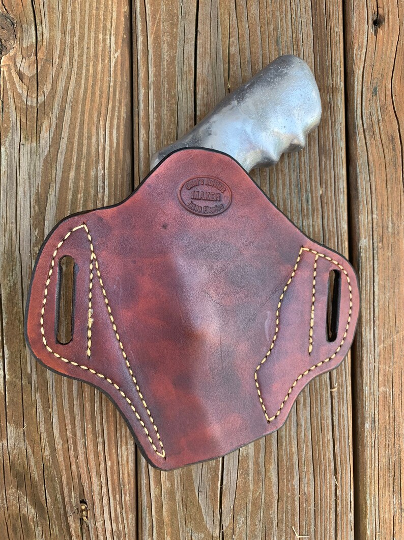 May include: A brown leather holster with a light brown stitching detail. The holster has a cut-out for a gun and a maker's stamp that reads "Custom Leather Maker, Saddle Fitting".