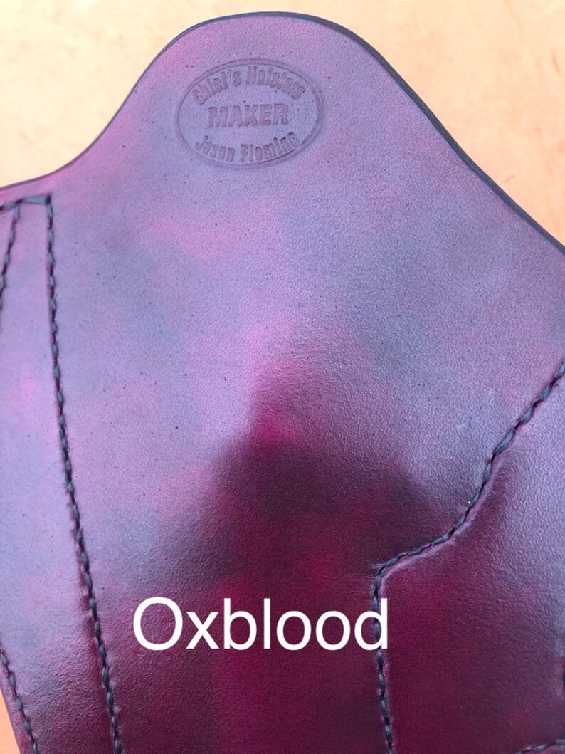 Pu&ograve; includere: Primo piano di una fondina in pelle rosso scuro con il testo "Glock's Holster Maker Jason Fleming" inciso in un cerchio. Il testo "Oxblood" &egrave; stampato sotto la fondina.