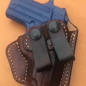 May include: A brown leather holster with two black leather straps and snaps. A blue handgun is inside the holster.