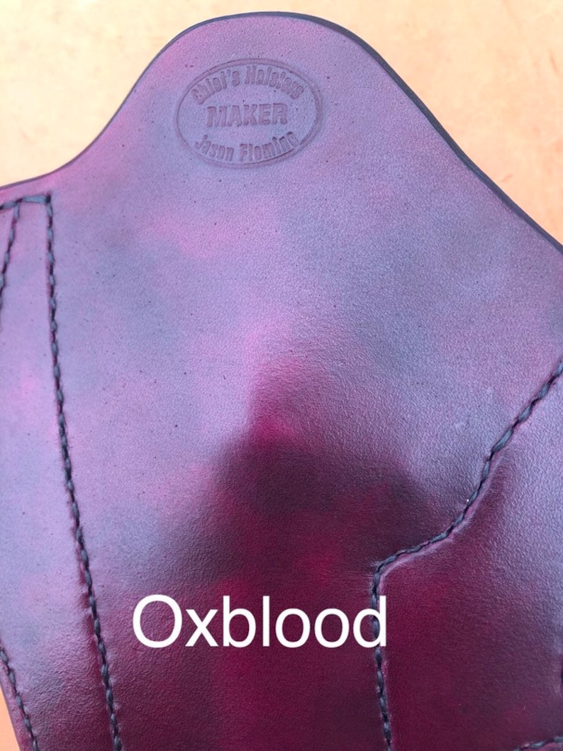 May include: A close-up of a dark red leather holster with the text "Glock's Holster Maker Jason Fleming" embossed in a circle. The text "Oxblood" is printed below the holster.