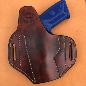 Pu&ograve; includere: Un fodero in pelle marrone con dettagli di cucitura marrone chiaro. Il fodero &egrave; progettato per contenere una pistola Ruger blu. Il fodero ha un marchio del produttore che recita "Bulldog Leather Maker, Weston, Florida".