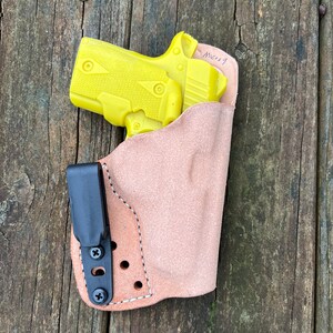 May include: A yellow handgun in a brown leather holster with a black clip. The holster has stitching around the edges and a few holes for attaching to a belt. The holster has the word "MCR19" written on it.