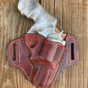 May include: A brown leather holster with a silver handgun inside. The holster has a stitched design and a belt loop.