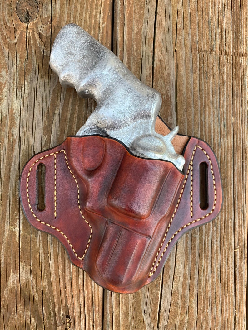 May include: A brown leather holster with a silver handgun inside. The holster has a stitched design and a belt loop.