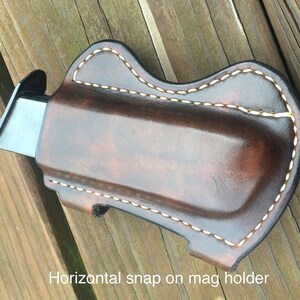 Pu&ograve; includere: Un portacaricatori in pelle marrone con cuciture bianche. Il portacaricatori ha una chiusura a scatto orizzontale. "Horizontal snap on mag holder" &egrave; scritto sotto il portacaricatori.