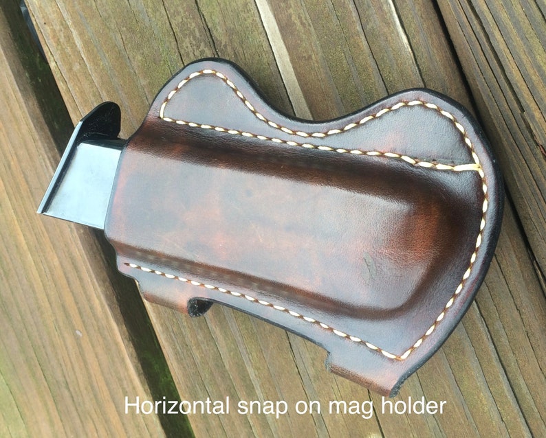 Pu&ograve; includere: Un portacaricatori in pelle marrone con cuciture bianche. Il portacaricatori ha una chiusura a scatto orizzontale. "Horizontal snap on mag holder" &egrave; scritto sotto il portacaricatori.