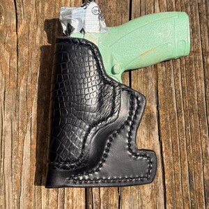 May include: A black leather holster with a textured crocodile-embossed pattern and visible stitching, containing a light mint green handgun with a small white and black label partially visible on the grip area of the firearm.