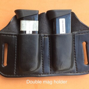 Pu&ograve; includere: Un portacaricatori doppio in pelle nera con due tasche per caricatori nere. Il testo "Double mag holder" &egrave; stampato sotto il portacaricatori.