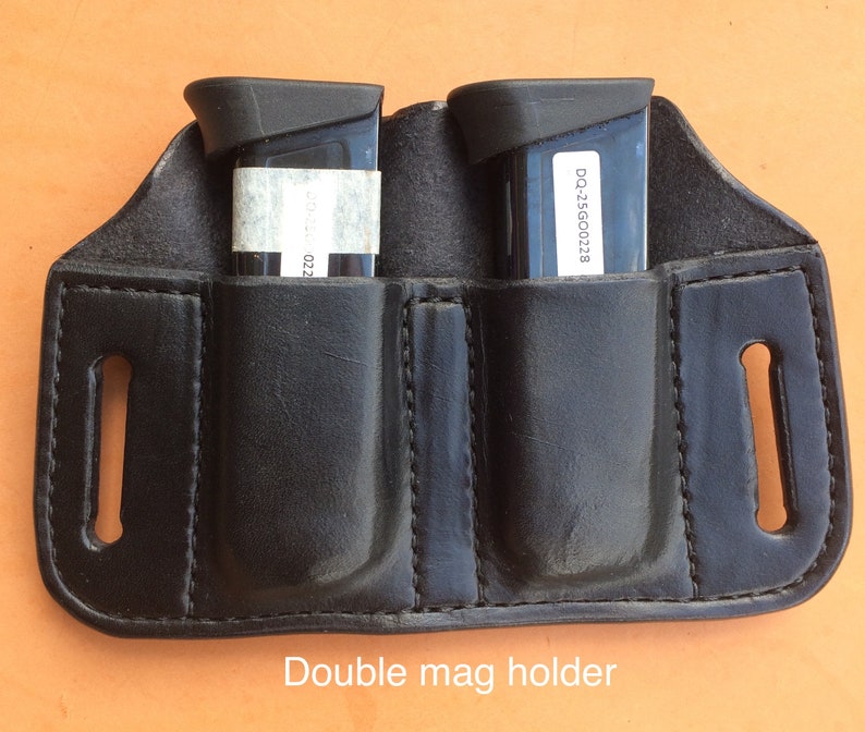 Pu&ograve; includere: Un portacaricatori doppio in pelle nera con due tasche per caricatori nere. Il testo "Double mag holder" &egrave; stampato sotto il portacaricatori.